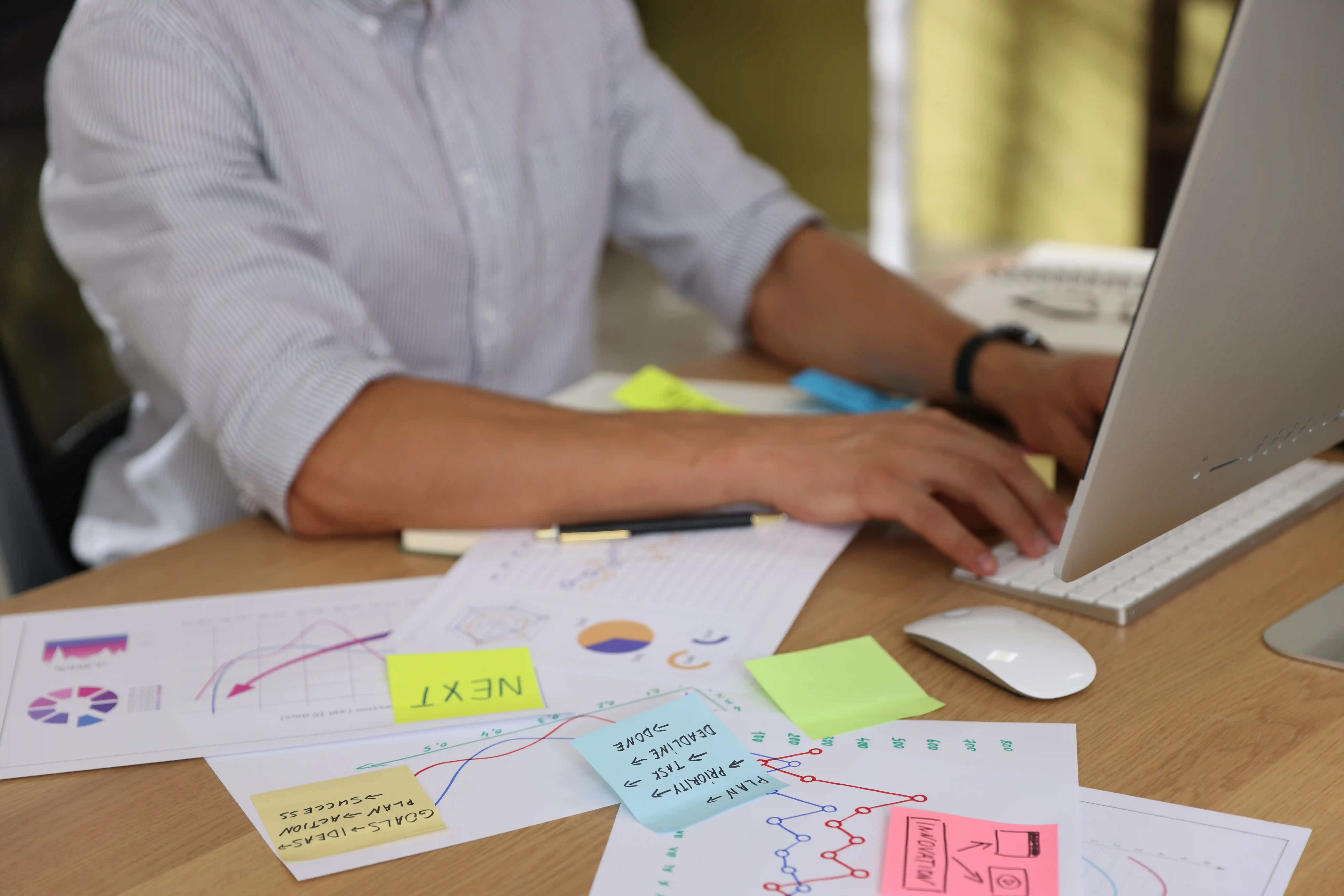 Person typing on a laptop at a desk covered with colorful sticky notes and printed charts.
